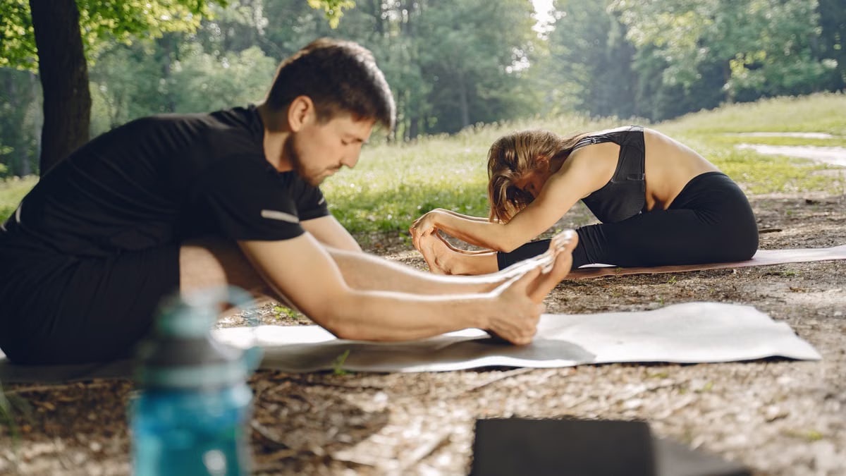 A man and woman practicing yoga outdoors in a sunny park, focusing on stretching.